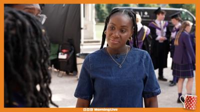 Presenter Tee Cee in the foreground wearing a blue denim dress, with Boarders actors in the background