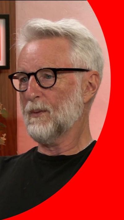 Billy Bragg has short white hair and a beard. He wears black glasses and a black top