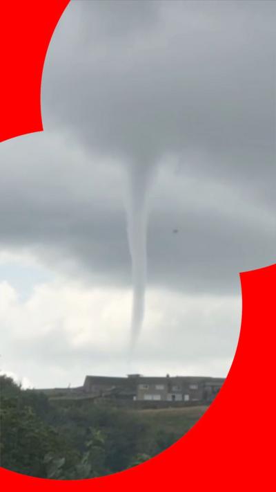 A tornado rises from a above a two-storey building in the distance. In the foreground is a field and vegetation.