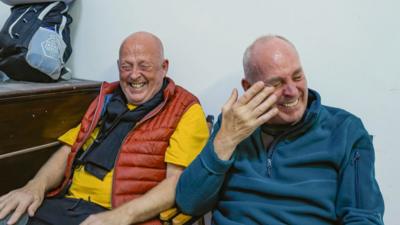 Two older men sitting beside each other laughing. One wearing red and yellow, the other in a blue jumper.
