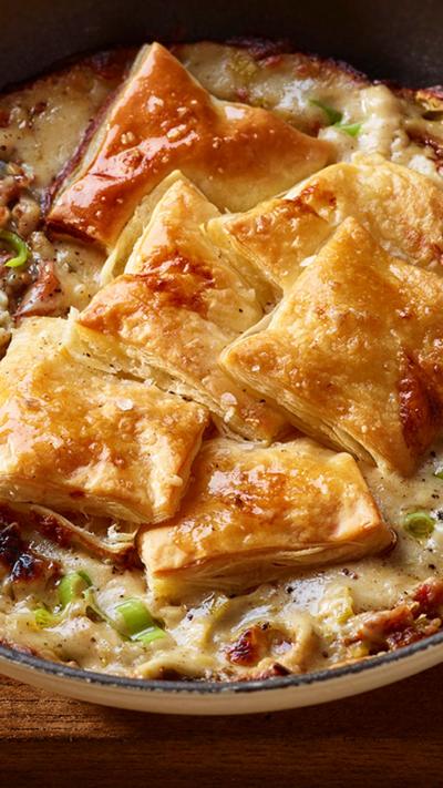A one-handled pan containing chicken, leek and bacon pie. A spoon sits in the side of the dish.