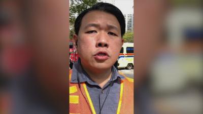 A man wearing an orange jacket over a shirt, with an ambulance and fire engine behind.