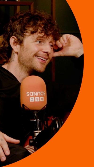 A man, Tom Grennan, smiling in a podcast studio, wearing black.