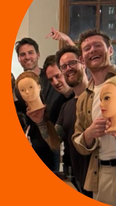 Four men smiling at the camera and gesturing to plastic heads with plaited hair.