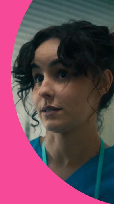 A woman with dark curly hair in a bun wears blue scrubs and has a lanyard round her neck