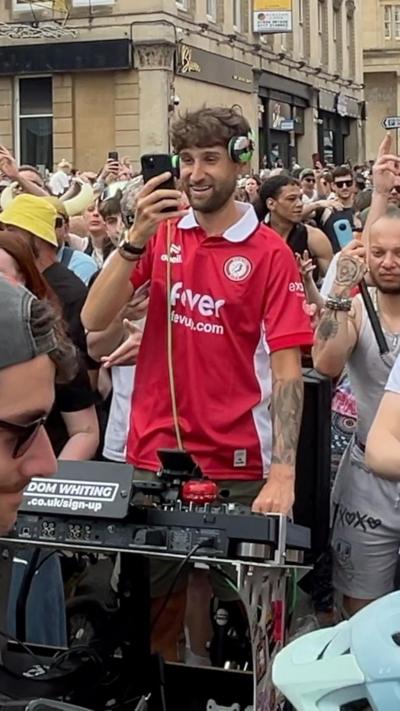DJ Dom Whiting, wearing a red Bristol City top, on his bike with DJ equipment in front of him. Behind is a large crowd of people.