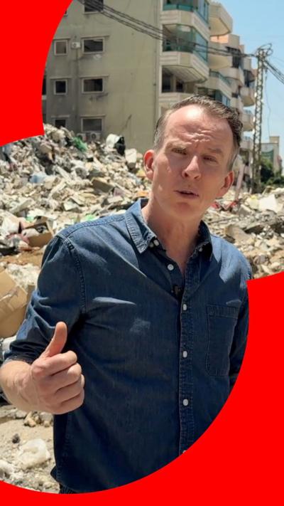 A man stands in front of rubble with a buildig in the background. He is wearing a blue shirt