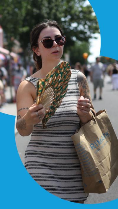 A woman with dark hair fans herself to keep cool. She is wearing sunglasses and a white dress with horizontal grey stripes. She is holding a paper bag.