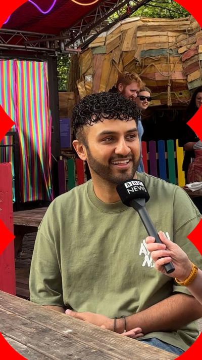 Shabaz Ali is a man with short black hair and a stubble beard. He is talking into a mic and laughing