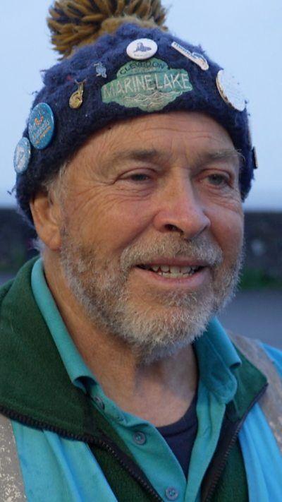 A man stood in a wool hat, stood outside, looking towards camera