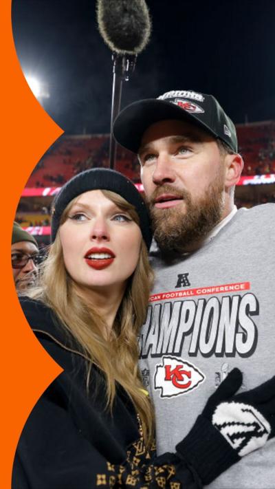 Taylor Swift and Travis Kelce embrace in a stadium. Both wear hats, Travis wears a grey T-shirt with 'Champions' on it