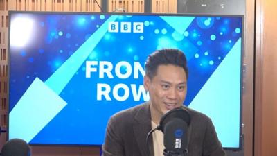 Jon M Chu speaking into a microphone in front of a large screen which says BBC Front Row