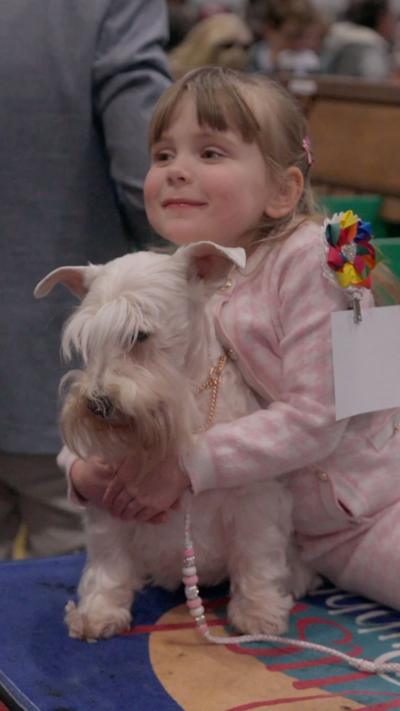 A child in a pink jacket with a fringe holding a white dog with a beard