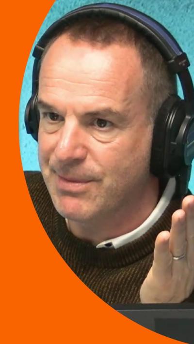 Martin Lewis wearing headphones in a podcast studio. He has short brown hair and is holding out his left hand
