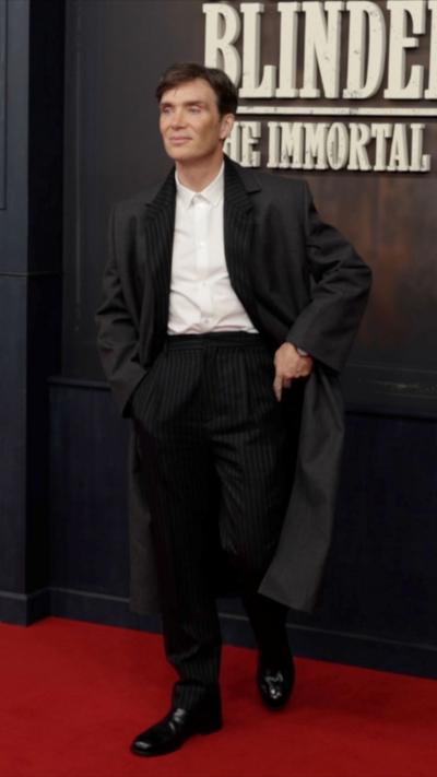 Cillian Murphy stood with his hands in his pockets wearing a long black blazer with a white shirt and striped black trousers