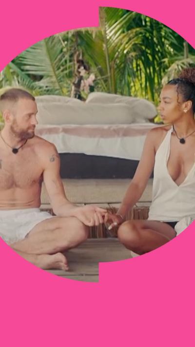A man and a woman sit cross legged holding hands. They are dressed in beach wear