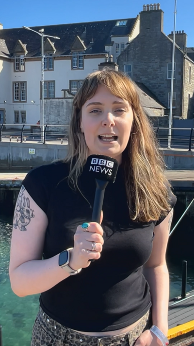Woman with long hair and a BBC News microphone standing on a quayside.