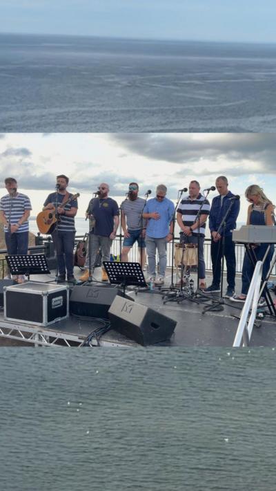 A group of eight performers stand on a stage outside singing into microphones. The view of the sea is behind them. One performer holds an acoustic guitar while another stands at a keyboard. 