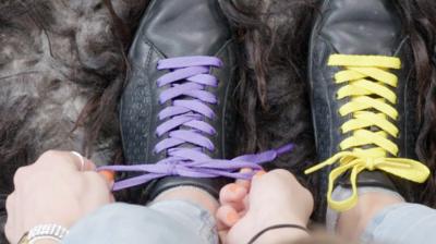 A pair of hands tying purple laces on a black shoe, beside the other shoe tied with yellow laces