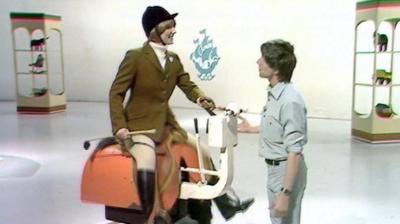 Lesley Judd sits on a mechanical horse. John Noakes stands beside her