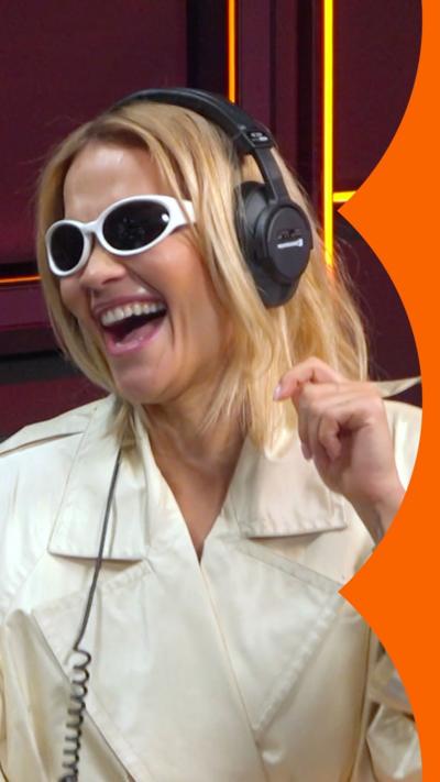 A woman, Rita Ora, with long blonde hair wears white sunglasses, a light coloured jacket and a pair of headphones. She is laughing.