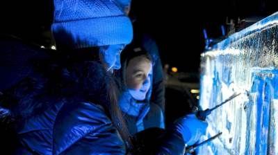 An adult female and young person hold chisels against a block of ice which is lit up with blue light. It reflects on their faces. They wear padded jackets, hats and scarves.