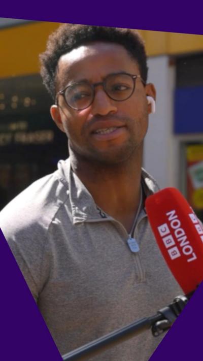 A young man wearing glasses stands in front of a BBC London mic on the street in Croydon.