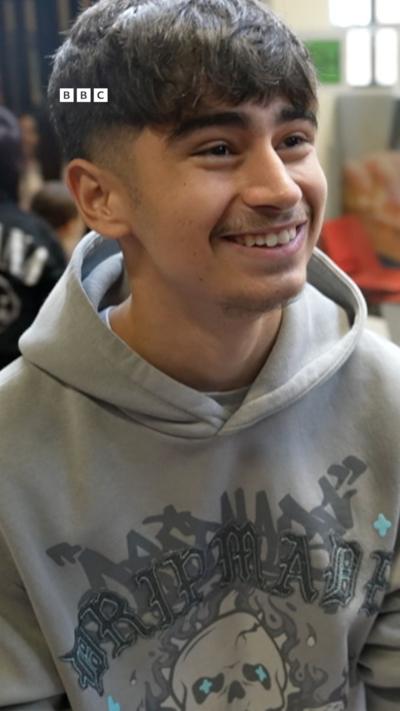 Boy smiling off camera, wearing a grey hoodie, moustache on his face, dark eyebrows and dark hair.