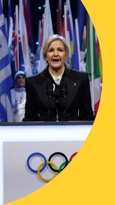 A woman with blonde hair and wearing a black jacket stands behind a podium bearing the Olympic rings on the front.