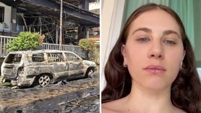 Splitscreen. Left, a burnt out car. Right, a woman speaks to camera.