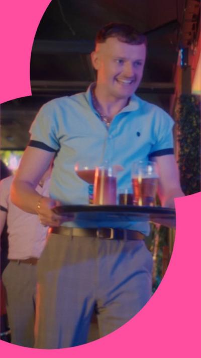 A man in a blue poloshirt carries a tray of drinks through a bar