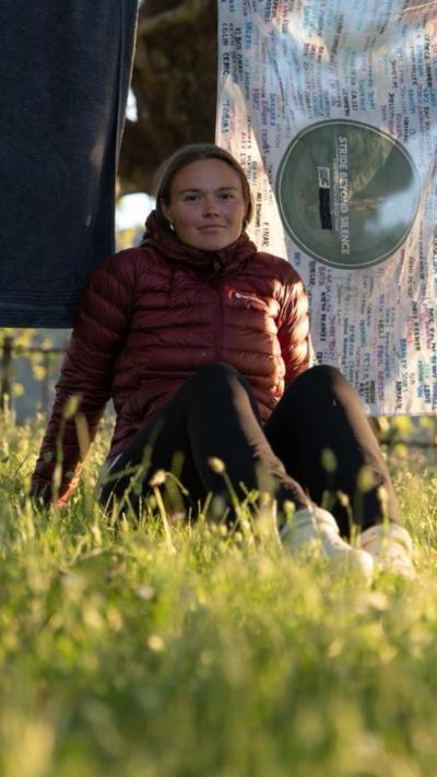Kit sits in the grass outside. She wears a maroon down jacket and black trousers, has brown hair tied back in a low ponytail and there are blankets hanging on a wash line behind her.