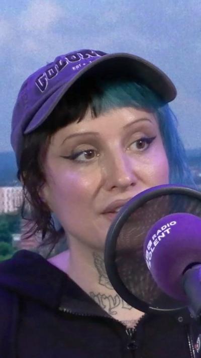 Rox Pink sits in the Radio Solent studio with a blue background. She has her hair tied back in a ponytail with a fringe. Half her hair is died blue and half is black. She has a black peaked cap on her head and tattoos on her neck and wears a black zipped up hoodie.