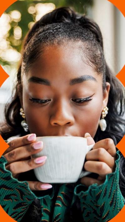A woman with black hair tied back, looks down to a white cup in front of her mouth, held with both hands.