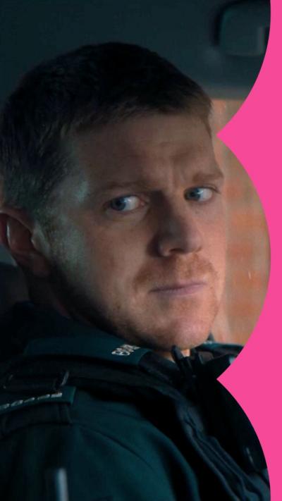 A uniformed police officer in a car, looking wearily to his right. He has short ginger hair and stubble