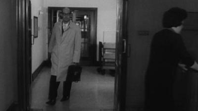 Black and white image of Philip Larkin in a long white coat, carrying a briefcase, walking through a library door.  In the foreground a women pushes a trolly of books.