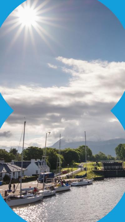 The sun shines in a cloudy blue sky down on a canal where several sailing boats are moored up. There are mountains in the background.