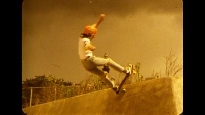 A boy on a skateboard with a white jumper and orange helmet in washed out 1980s film footage.