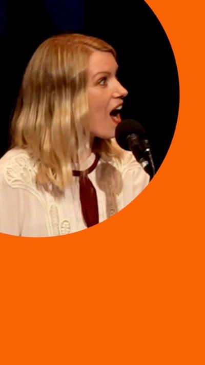 Rachel Parris, a woman with blonde hair and a white shirt, sits in front of mic, open mouthed in shock.