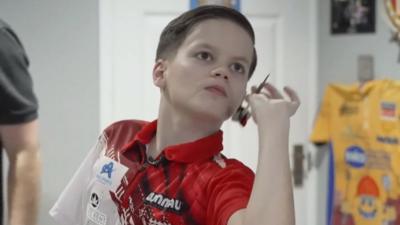 Jack Johnson, a young white teenager wearing a red and white darts shirt aims a dart at a dart board. 