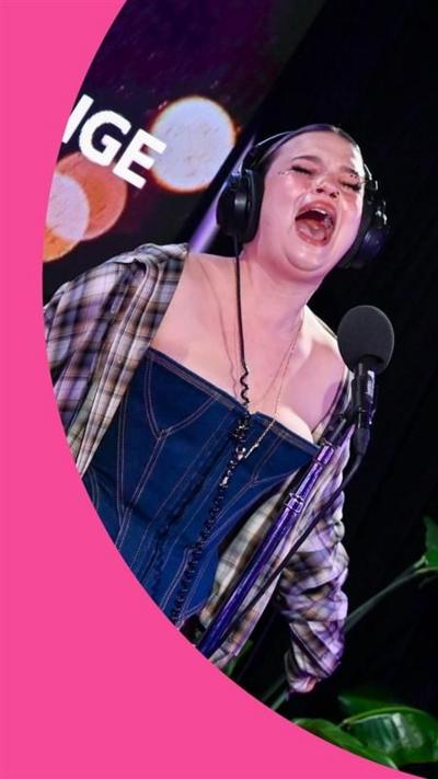 Lola Young sings in the Radio 1 Live Lounge studio. She wears a checked shirt and trousers, and a denim corset