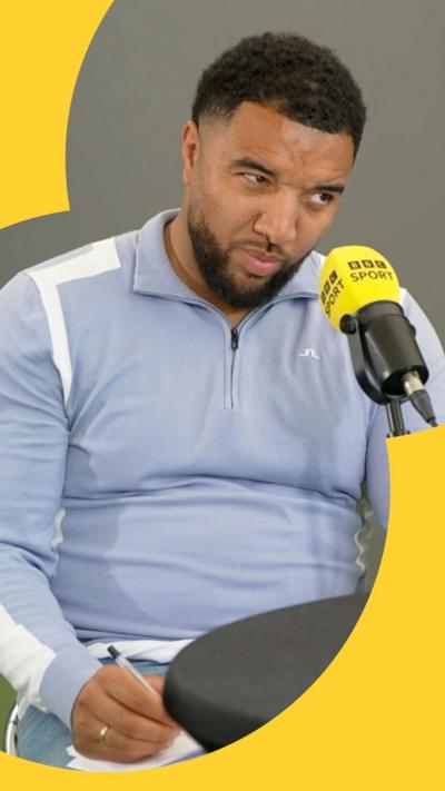 A man, Troy Deeney, looks judgingly sat behind a BBC Sport mic