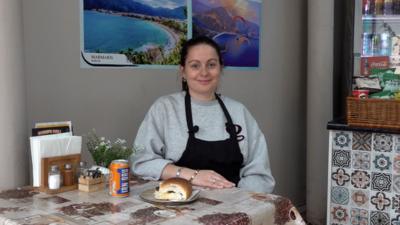 A women with brown hair and blue eyes wearing a green jumper under a black apron sits with a lorne sausage roll and Irn Bru at a table in a cafe.