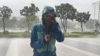 BBC correspondent Laura Bicker, wearing a blue jacket, shields her face from wind and rain. Behind her, wind blows trees against a grey sky thick with cloud. 