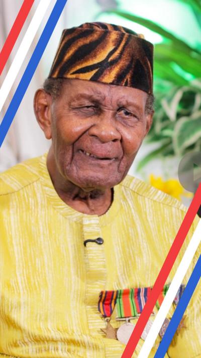 100-year-old Ghanian veteran Joseph Hammond sits reflectively beside his autobiography
book on a table with potted plants behind him, while speaking to a reporter in an interview about his time fighting in Burma. He is wearing a black and brown striped hat and a yellow and white striped top with five war medals shaped as circles and stars pinned to it.