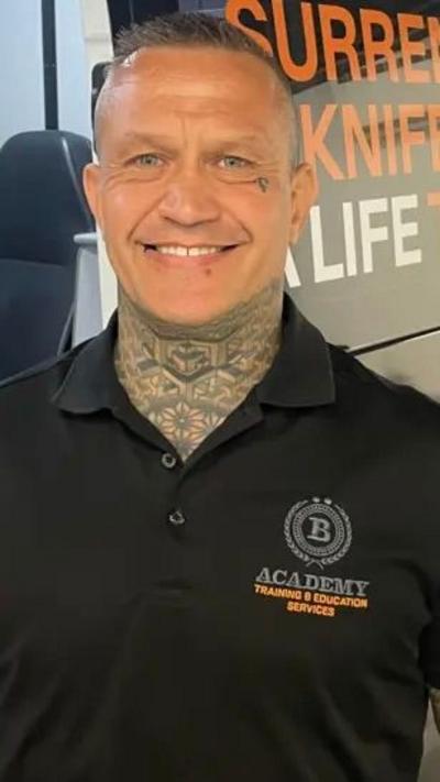 A man with short hair, a black shirt and tattoos on his arms and neck, smiles as he stands in front of a van with branding on it and a chairs, table and laptop inside.