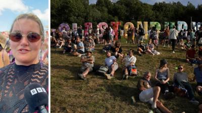 Woman wearing sunglasses on left side. Crowds gathered in front of Glastonbury sign
