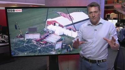 Joe Inwood stands in front of TV screen showing picture of stage with seats lying on the floor in front