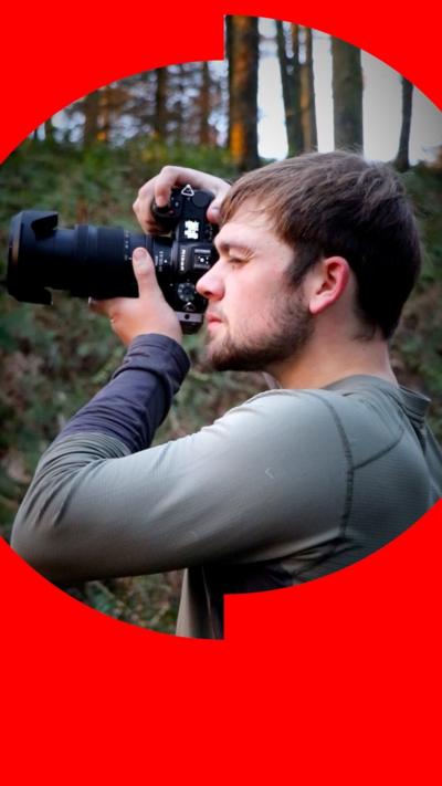 A young man wearing a grey jacket and black rucksack stood in front of a lake and a hill