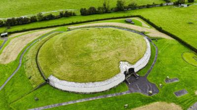 Picture of an ancient burial tomb - it is photographed from above, the tomb is round with a stone wall and grass growing on its roof. It is surrounded by grass and fields. 